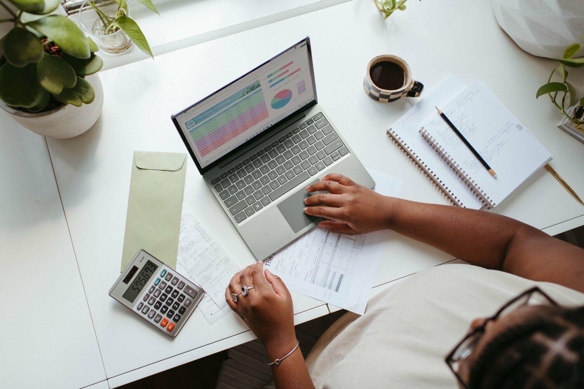 Person using a laptop in a home office with plants, coffee, and office supplies around.