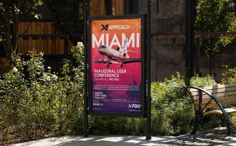 Outdoor sign promoting Miami user conference with an airplane graphic, surrounded by lush greenery.