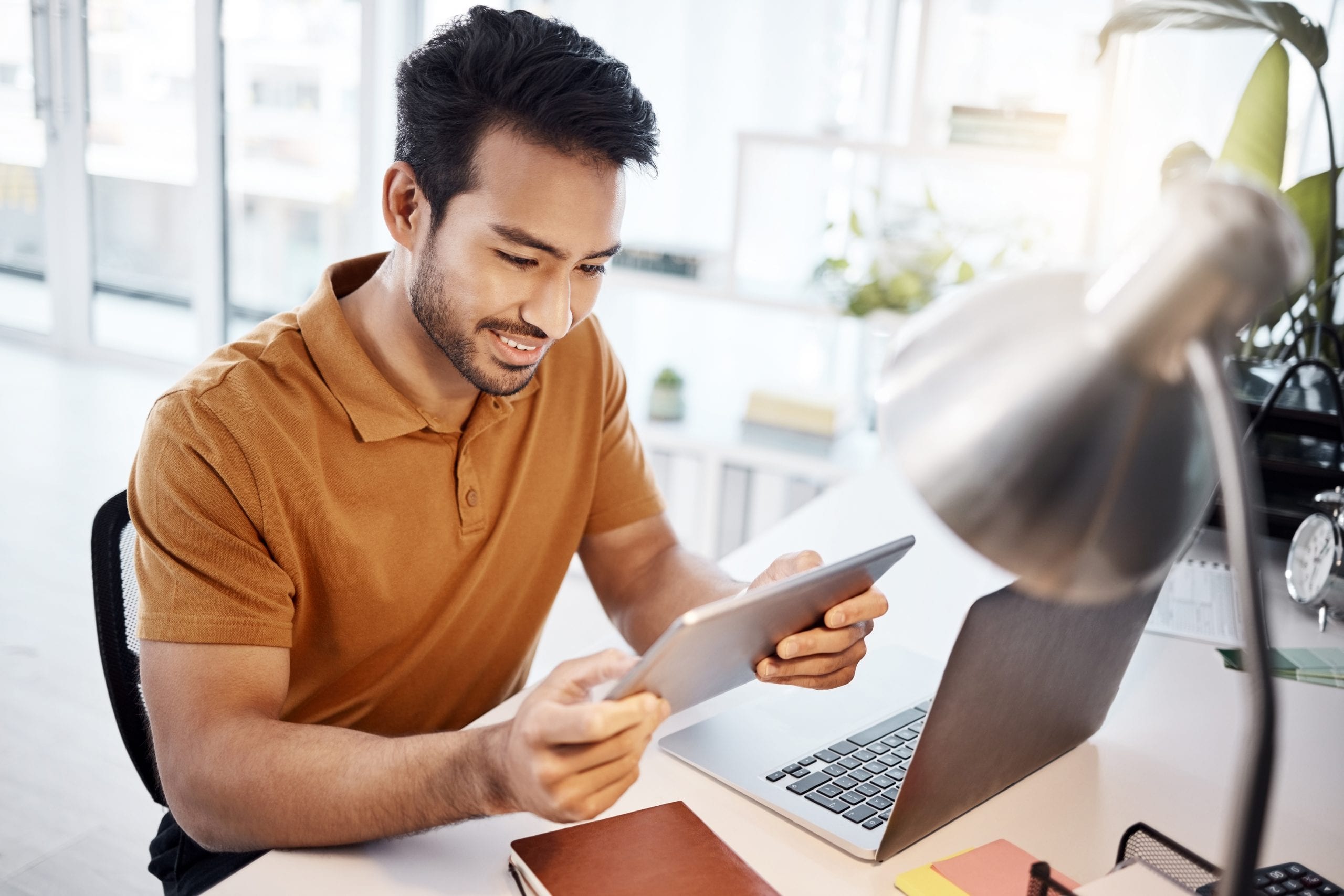 Man smiling while reading on a tablet at a modern office desk with a laptop and notebooks.