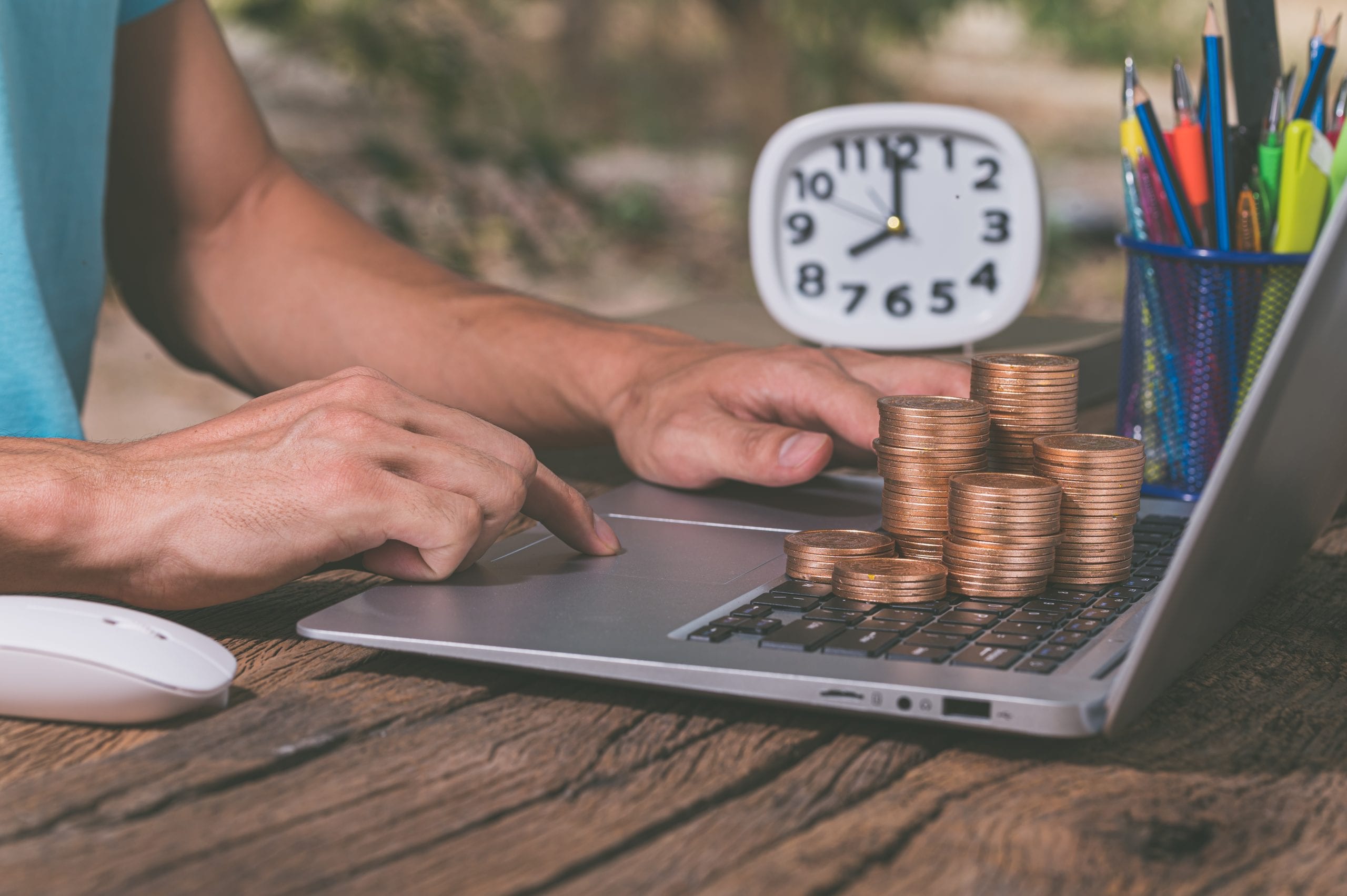 Person typing on a laptop surrounded by stacked coins and a clock, symbolizing online work and financial growth.