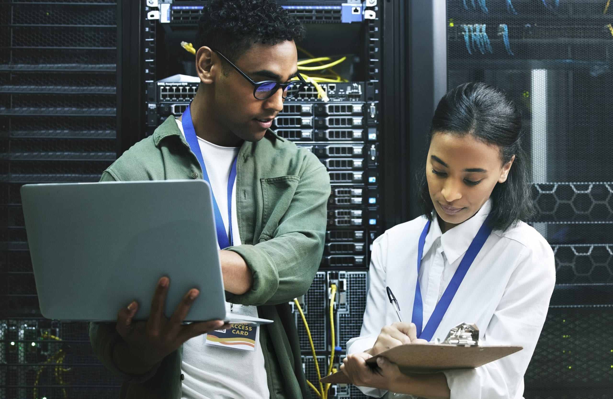 Two technicians checking servers; one uses a laptop while the other writes on a clipboard in a server room.