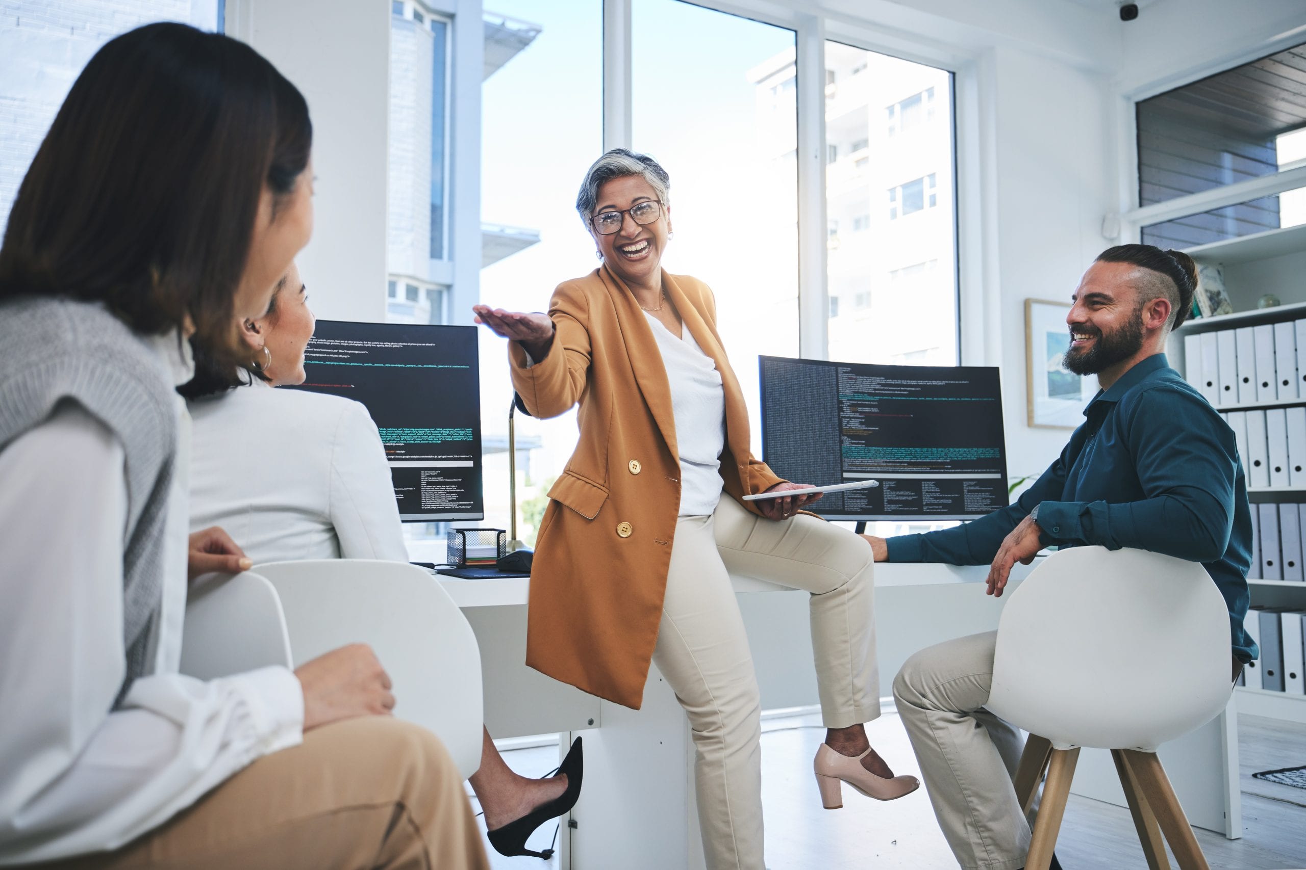 Diverse team in a modern office engaged in a collaborative meeting with a mentor, two monitors displaying code.
