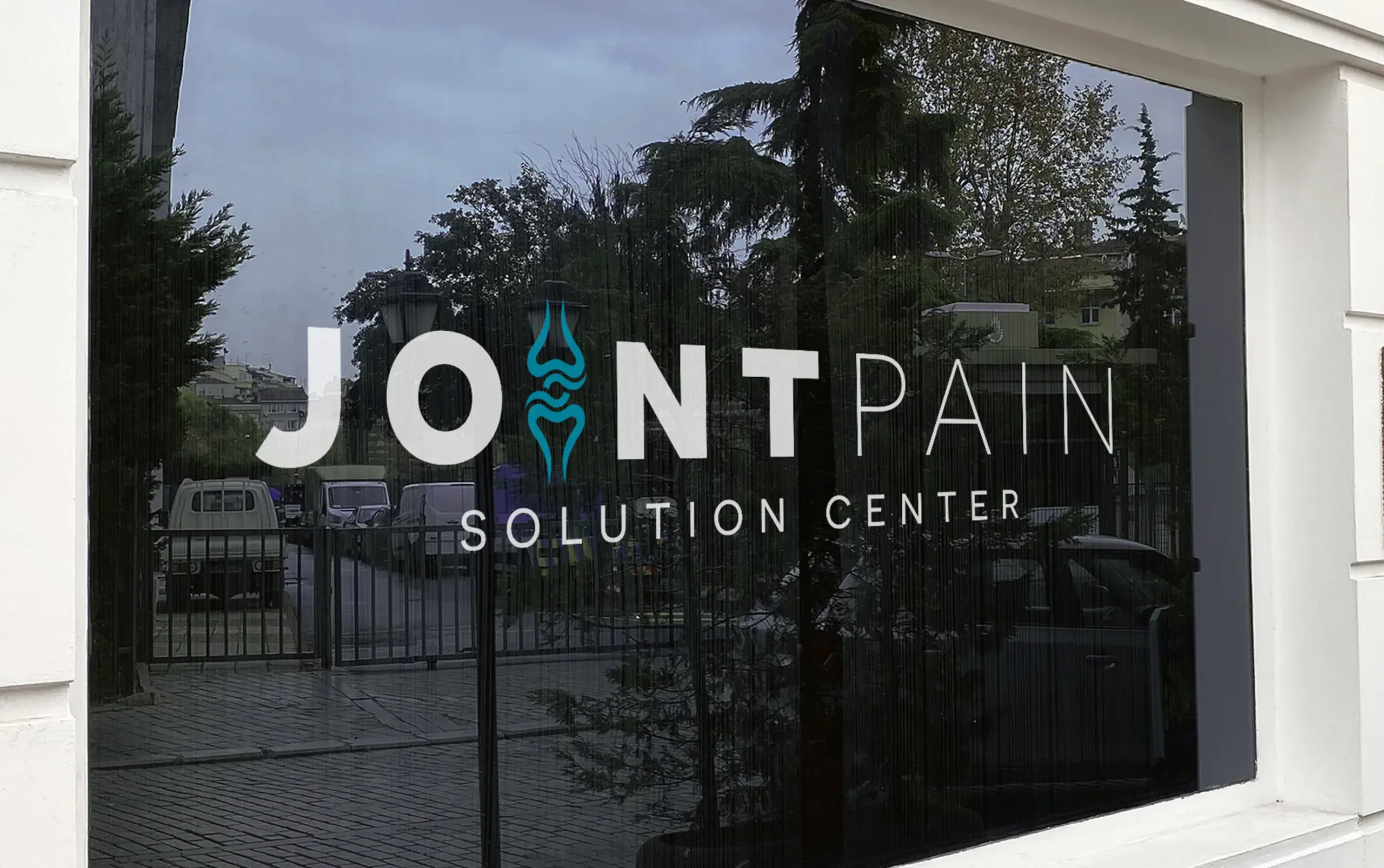 Sign for Joint Pain Solution Center displayed on a glass window with trees and buildings in the background.