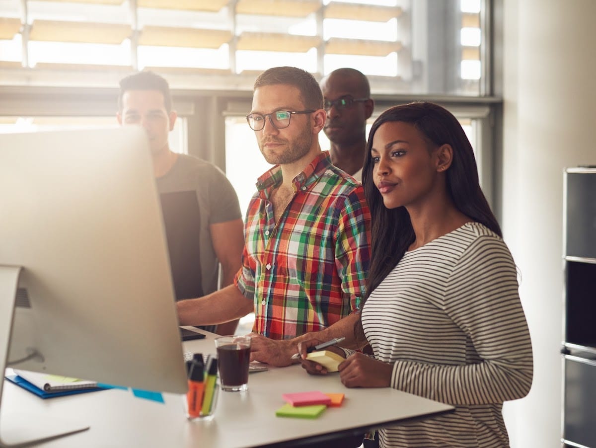 Four diverse workers collaborating at a computer in a modern office setting.