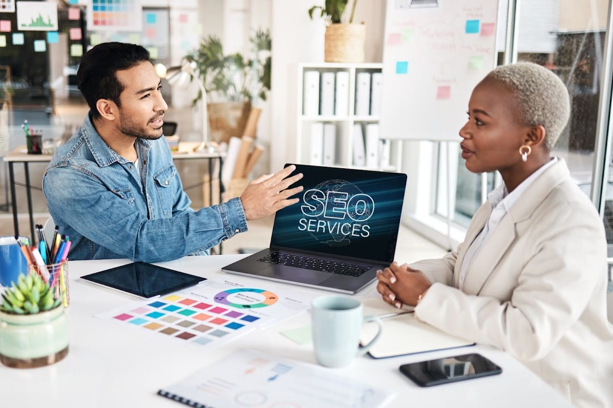 A young professional presenting SEO services on a laptop to a colleague during a meeting. The setting is a modern office space, and the discussion appears focused on strategies to enhance online visibility.