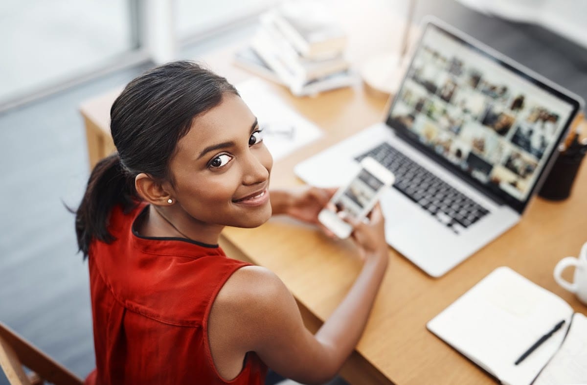 Woman smiling at desk with laptop and smartphone, surrounded by office supplies.