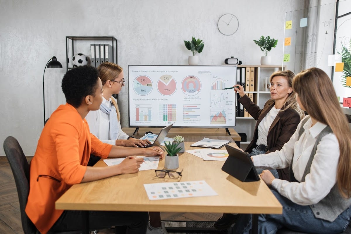 Diverse businesswomen collaborate around a desk, discussing strategies with charts displayed on a screen.