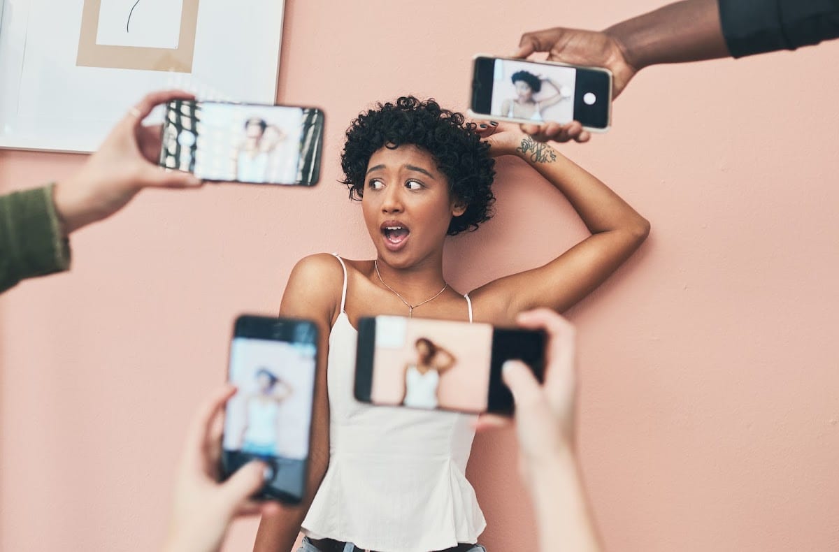 Young woman posing with surprised expression surrounded by phones capturing her image against a peach background.