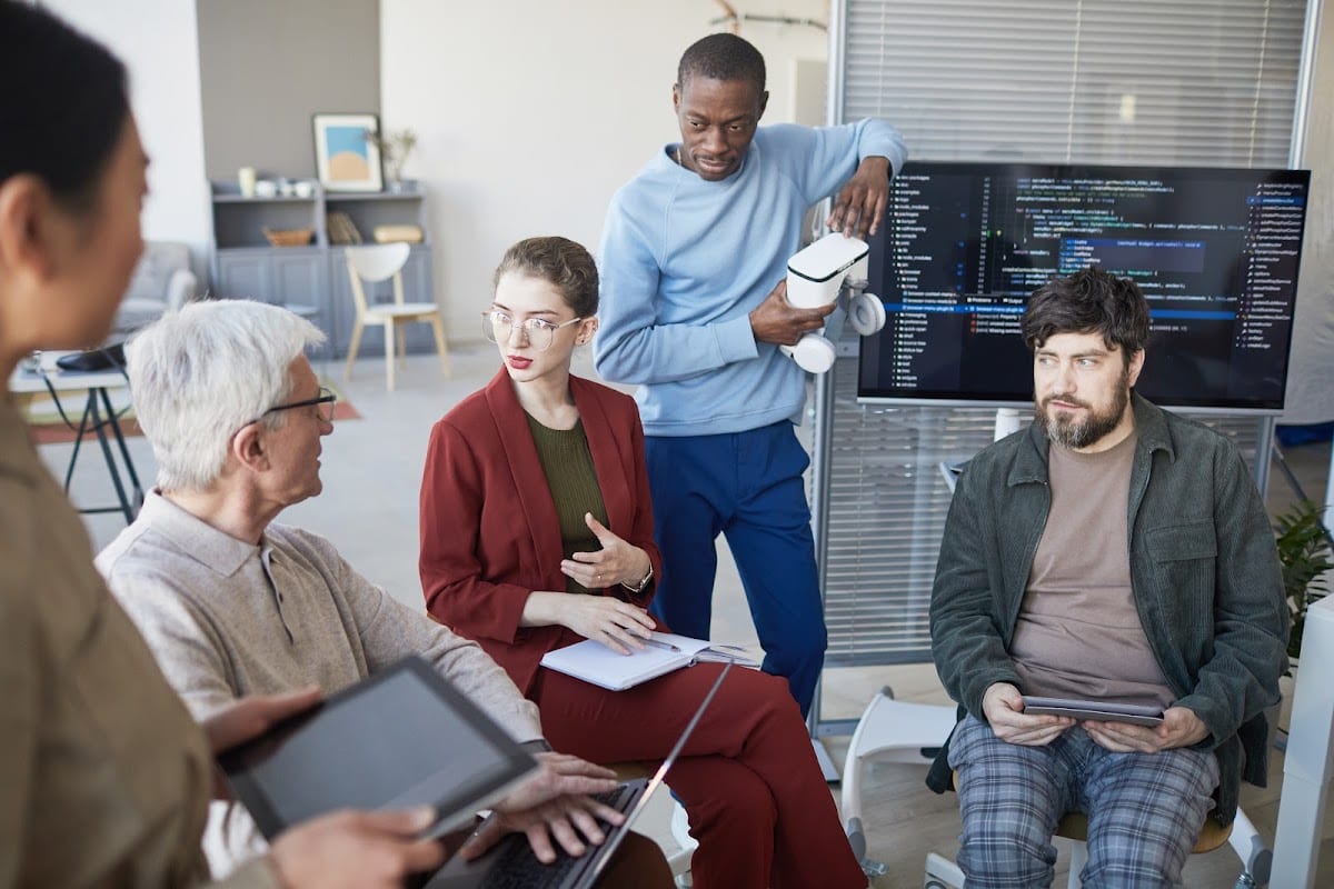A diverse team of software developers and engineers collaborating in a modern office with a screen displaying lines of code. The team discusses software development strategies with a mix of laptops, tablets, and VR equipment.