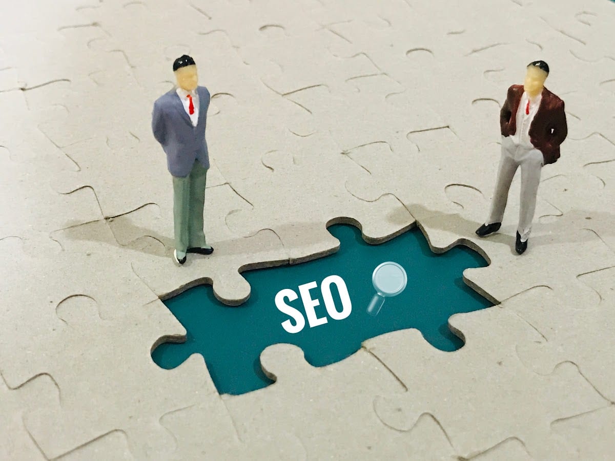 Two miniature figures standing on puzzle pieces revealing the word SEO.