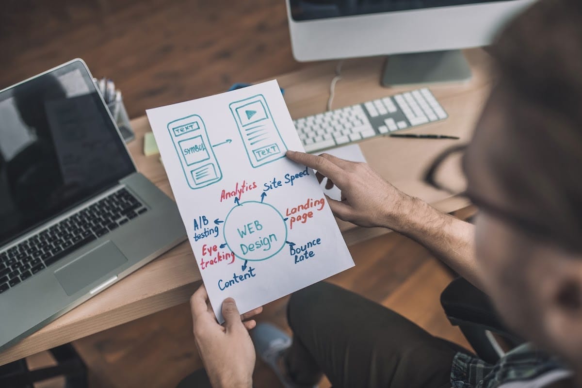 Man in glasses holding a web design draft at a desk with a laptop.